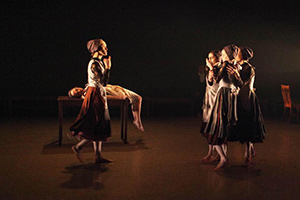 A woman walking toward a group of three women huddled together. There is somebody laying on a table in the background.