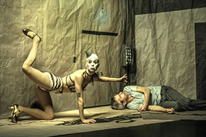 A woman in clown makeup kneeling in an odd pose looking at the camera. There is a man in the corner of the room laying on his side.