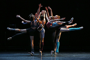 Several woman in a large dark space huddled together facing each other, all of which are in a ballerina pose standing on their toes with one leg stretched out and arms up.