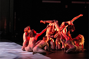 Several woman under a red light. Most of them are sitting on the ground holding one of the women up, who is leaning back with an arm and a leg raised. There is a large square beam of light to the left of them.