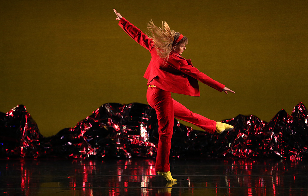 A woman dressed in red posing with arms spread out and pointing one leg out.