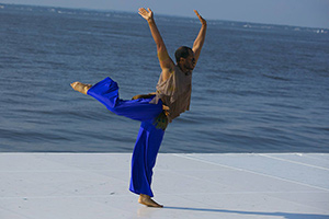 A man in blue pants standing on one foot with arms up in front of the ocean.