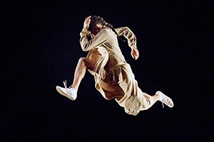 A woman in a beige dress jumping in the air, in a "running man" pose.