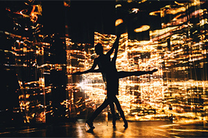 The shadow outlines of two people dancing with arms and legs spread out in front fo yellow lighting.