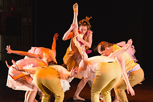 Six dancers in yellow costumes bent over in a circle with one dancer in the middle standing with one leg held high over her head.