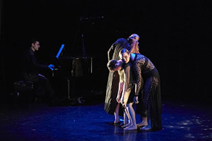 Dancers on stage huddled together and bending at the waist with black background.