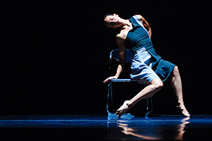 A woman on stage on the edge of a chair, one are supporting her, bare feet with points toes. Blue dress and lighting.