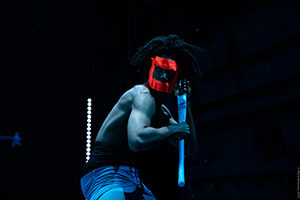 A man on stage with bare chest and red mask holding a club with both hands.