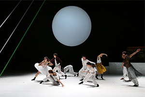 Nine dancers on a stage with a black background and large white sphere in middle. Dancers are onn in different poses and dressed in mostly white.