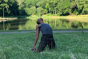 Man kneeling and leaning backwards in grass with pond and trees in background.